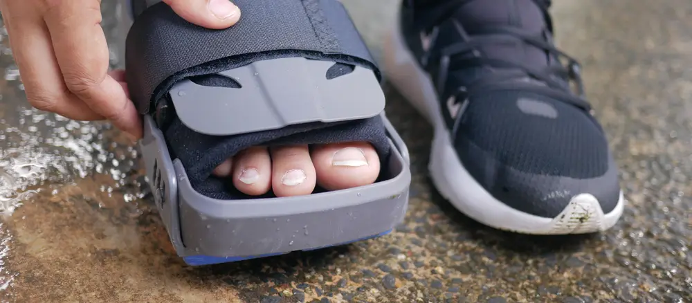 A hand fastening the front strap of a grey CAM walker boot on one foot, with a sports shoe on the other foot.